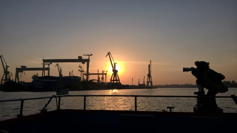 Silhouette einer Werftanlage in Mangalia bei Sonnenuntergang, im Vordergrund eine stilisierte Seefahrerfigur mit Fernrohr an einer Reling