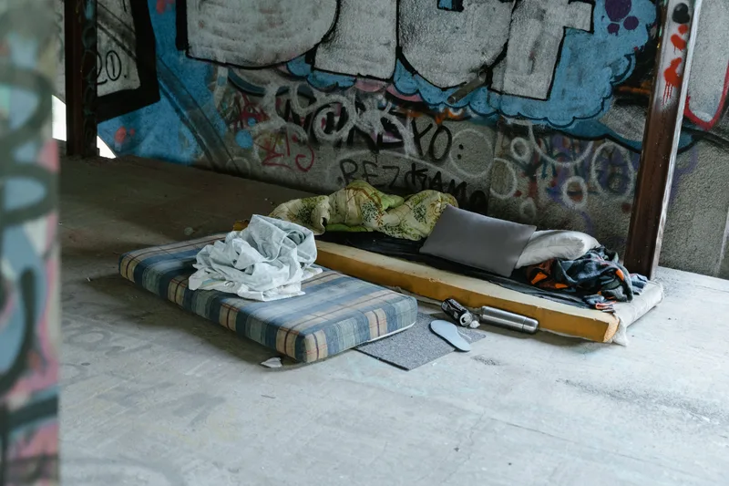 Behelfsmäßiges Schlaflager mit Matratzen und persönlichen Gegenständen auf dem Betonboden eines mit Graffiti verzierten Rohbaus. Gefahr im Winter