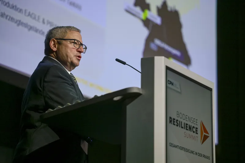 Referent am Rednerpult beim Bodensee Resilience Summit vor einer Präsentation über die Fertigung von Eagle- und Piranha-Radfahrzeugen