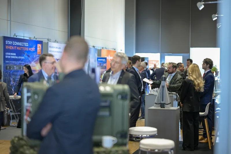 Fachbesucher im Gespräch auf einer Rüstungsmesse vor Diehl-Exponaten, Satellitenkommunikationstechnik und militärischen Transportbehältern