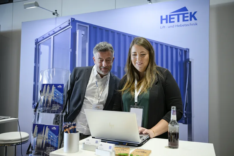 Zwei Personen arbeiten an einem Laptop am Messestand der Firma HETEK Lift- und Hebetechnik vor einer Grafik eines Container-Hebesystems