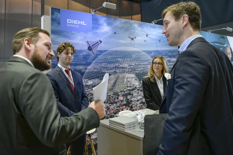Fachbesucher im Gespräch am Messestand von Diehl Defence vor einer Schautafel mit Drohnenabwehrsystemen