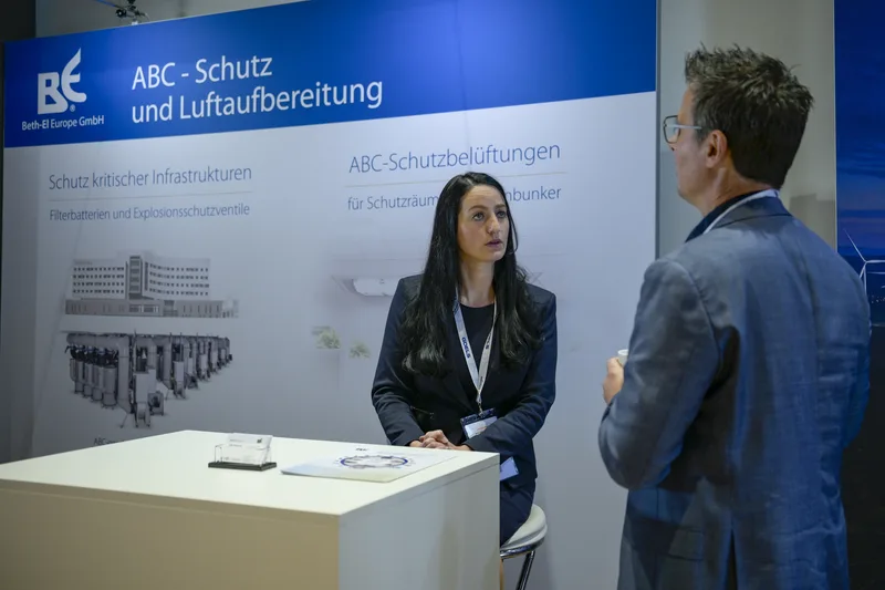 Unternehmensvertreterin der Beth-El Europe GmbH im Fachgespräch mit einem Besucher an einem Messestand für ABC-Schutzsysteme