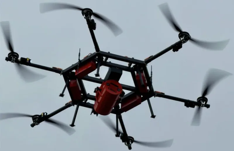 Hexakopter mit sechs Rotoren, sechseckigem Rahmen, roten Akkupaketen und zentraler zylindrischer Nutzlast im Flug vor hellem Himmel