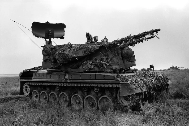 Flugabwehrkanonenpanzer Gepard der Bundeswehr mit getarnter Besatzung im Gelände und einem weiteren Militärfahrzeug im Hintergrund