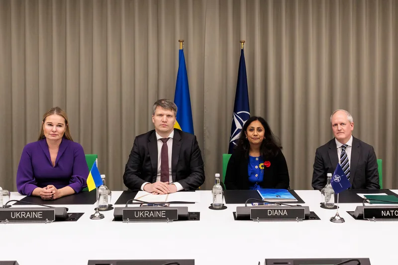 Vertreter der Ukraine, der NATO und von DIANA sitzen bei einer Konferenz an einem Tisch vor den Flaggen der Ukraine und der NATO