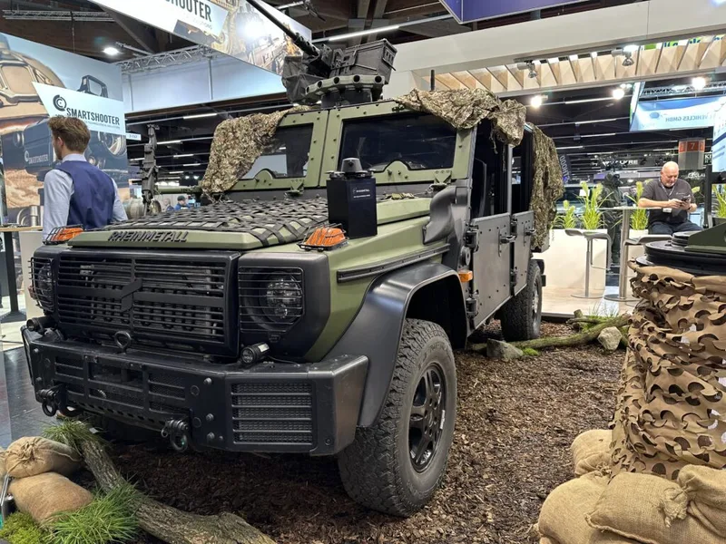 Rheinmetall Caracal Einsatzfahrzeug mit Dachwaffenstation und Tarnnetz auf einem Messestand mit Sandsäcken und Naturmaterialien